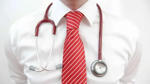 PA Media A close up of a man's chest. He is wearing a white shirt and red tie and has a red and silver stethoscope hanging around his neck.