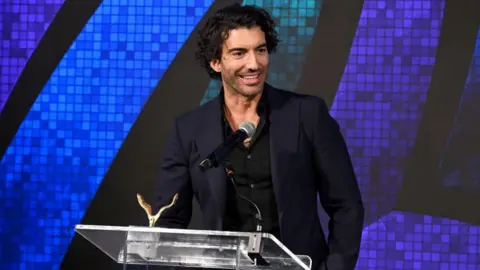 Actor Justin Baldoni is seen on a stage accepting an award in New York. He is wearing a dark-coloured suit and is standing at a microphone with a golden award sitting on the lectern. 