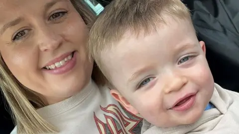 Lydia Collins with fair shoulder-length hair, smiling at the camera while holding her son Rylo, aged about two, with fair short hair and blue eyes' He is also smiling