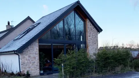 The holiday let building, a large window the entire height of the wall is visible. Pine trees are planted in front of it.