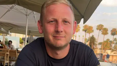 Jordan Hattersley, a man with blond hair and blue eyes and a dark t-shirt. He is smiling into the camera. In the background are parasols and palm trees.