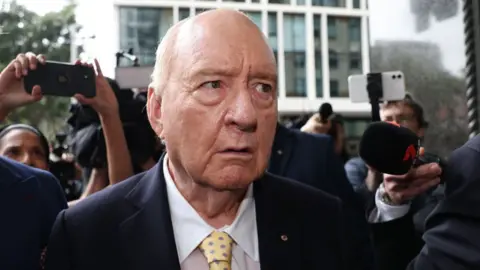 Alan Jones wearing a black suit jacket, white shirt and yellow tie arriving at court