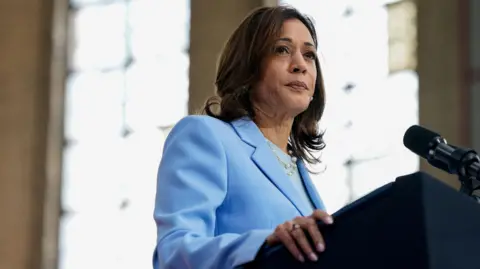 Kamala Harris wears a blue suit and looks out of frame while standing at a microphone