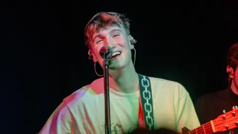 Singer Calum Bowie on stage performing, smiling and holding guitar 