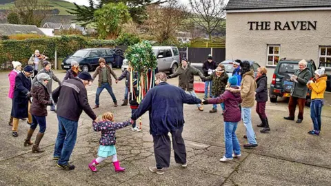 A group of people holding hands in a circle and dancing in a pub car park, with a building that has a sign reading The Raven on the wall in the background, along with hills and trees. 