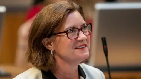 A head a shoulders picture of Eluned Morgan wearing glasses and smiling in the Senedd chamber with a microphone in the background