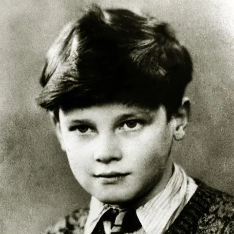 Alamy A black and white image of Sir Roger Moore as a child. He has a serious but pensive expression on his face and is wearing a stripy shirt, a tie and a patterned knitted jumper. He has a full head of dark hair.