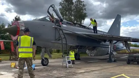 Several people, men and women, wearing hi-vis vests and camouflage clothing, carry out maintenance work on the Lightning jet. The plane is outside on a tarmacked runway area.