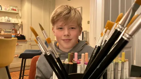 Reuben looks into the camera and is surrounded by pens, paint brushes and other art equipment. In the background a number of light coloured cupboards and drawers can be seen. There are a number of family photographs on a shelf too. Reuben has blond hair which is swept to one side and is wearing a grey hoodie.