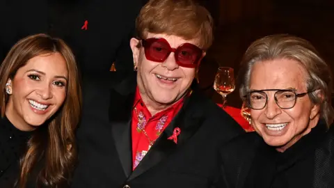 Elton John smiles while looking at the camera through purple sunglasses, wearing a red shirt and black suit, posing in between Patricia and Richard Caring, while attending the premiere on Sunday