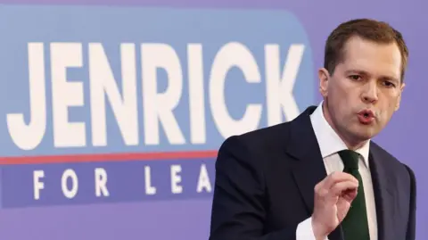 Robert Jenrick speaking at conference in front of a sign which states Jenrick for leader