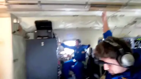 A man in a blue jumpsuit braces against the ceiling with another person in a blue jumpsuit behind him