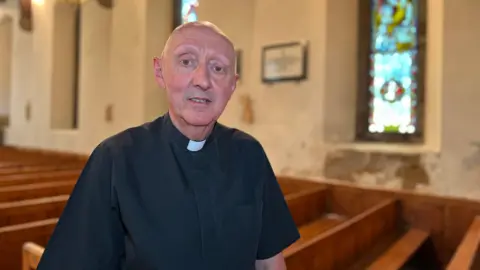 BBC Father Barry looks at the camera in front of a row of pews but in the background there is plaster falling off the wall under a window