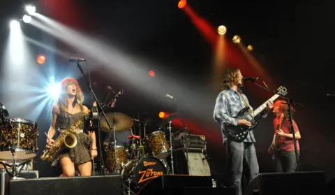 Zutons members Abbie Harding, Sean Payne and Dave McCabe perform on stage at the Cambridge Folk Festival in 2009. Abbie is playing saxophone, Sean the bass and Dave the guitar.