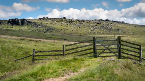 A view of a grassy moor on a sunny day with blue skies and clouds seen above a rocky outcrop, which gives way to a grassy hill with a fence and footpath seen in the foreground. 