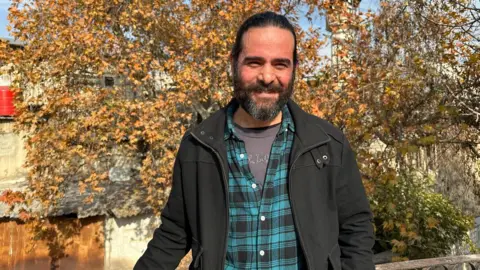 Wajd Khair, wearing a t-shirt, button-down and jacket, stands and smiles as he looks into the camera, a tree with yellow leaves behind him 