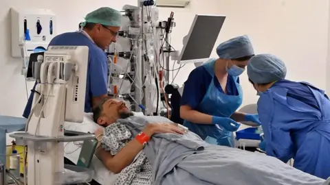 Royal Papworth Hospital Daniel Evans-Smith lying on a bed in the operating theatre at Royal Papworth hospital. He has his eyes open and a wristband on and is looking up at the ceiling. A surgeon stands behind him wearing a hair net and scrubs. Two nurses are checking equipment to his left and are wearing scrubs and aprons