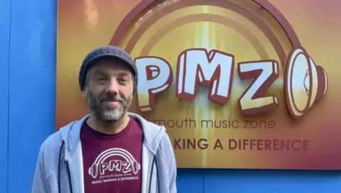 Karl Meyer, who runs Plymouth Music Zone, is pictured infront of an orange sign, which has the Plymouth Music Zone logo on it. The logo is a picture of animated headphones with PMZ in the middle. Below it it reads 'Plymouth Music Zone' and below that it reads 'Making a difference'. Karl is wearing a burgundy T-shirt with the same logo on it and a grey hoodie on top.