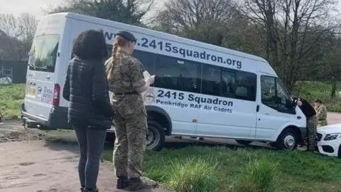 Two people are stood at the side of a minibus, one with a black padded jacket on and the other with a camouflage outfit, while two others are stood at the front of the vehicle, one of whom is in between the minibus and the front of a white car