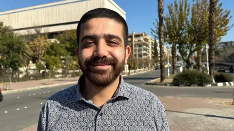 DJ and musician Maher Green, wearing a collared shirt, looks into the camera, with a building, road and trees behind him