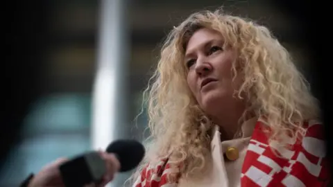 Getty Images Tessa Gorman speaking to press wearing a patterened red and white scarf and pink jacket with gold buttons