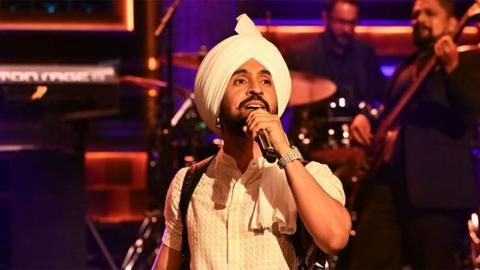 Diljit Dosanjh, an artist singing into a black microphone, wearing a cream coloured outfit and turban with a shiny watch on his left wrist. In the background you see two men out of focus playing an instrument.