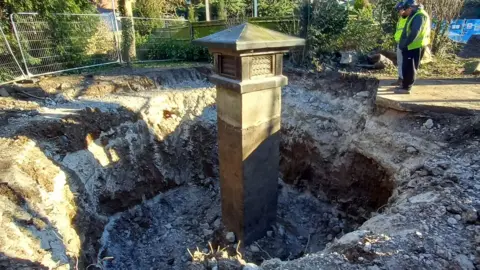 MOLA A dug-up area revealing a deep excavation with a WWII-era ventilation shaft emerging from the ground, surrounded by construction fencing and workers in high-visibility vests inspecting the site.