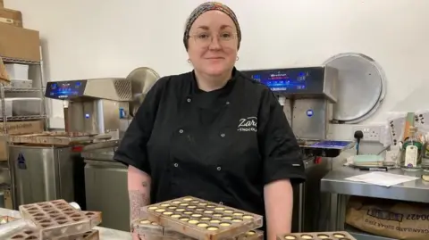 Helen Jones, head chocolatier at Zara's chocolates, wearing a head bandanna and a black top. She is standing in front of chocolate moulds on a desk, with mixing machinery and shelves behind her.