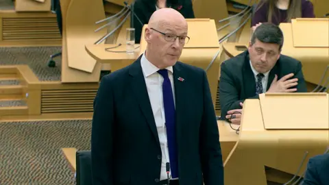 John Swinney standing in Holyrood's debating chamber. He is wearing a blue suit and dark tie and other MSPs can be seen behind him.