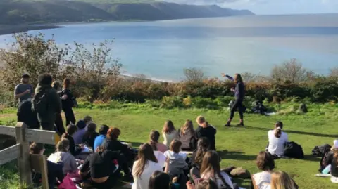 Image shows a group of young people sitting on a green verge that looks over Porlock Bay in Devon. Some of the teenagers are standing, others are sitting. A person is stood in front of the group and pointing out to sea, as if they are telling the group some information. 