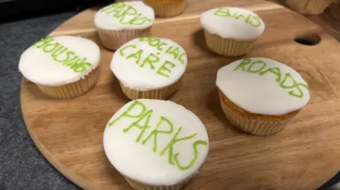 A batch of cupcakes iced with the names of council services, including parks, roads, social care, housing, bins and housing