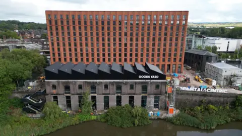 A red brick multi-storey building is surrounded by a building site. There is a smaller building in front that has a sign that reads Good Yards. With brown water in front of that.