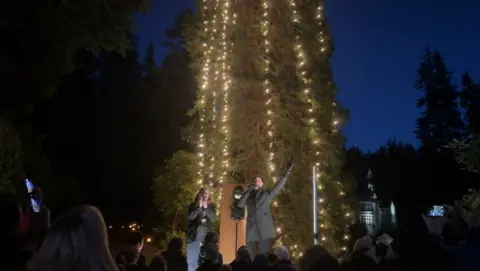 A tall tree is illuminated by thousands of yellow Christmas lights. George Clarke stands in front holding a microphone, with his other hand raised in the air. A crowd watches on.