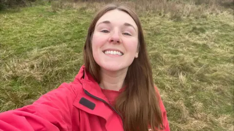 Becky Davies Picture of Becky Davies smiling in a field with her hair worn down behind her ears, wearing a pink raincoat.  