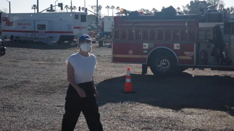 A woman wearing black trousers, a white t-shirt, a blue baseball cap and a white face mask is walking across gravel. Behind her is a fire engine and a white van marked "Mobile Kitchen Unit". 