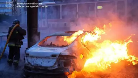 A firefighter works near a burning car with a damaged tram seen in the background after Russian shelling of Ukraine's southern city of Zaporizhzhia. Photo: 8 January 2025