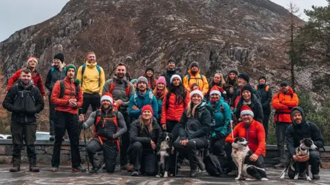 A group of hikers pictured infront of a mountain in Eryri National Park, people at the front are crouched down with dogs. 