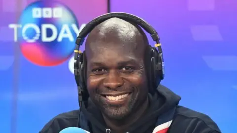 Chief scout Dwayne Fields in the Today studio. A light blue Radio 4 microphone can be seen in the foreground in front of him and the Today logo can be seen on screens in the background. Fields is wearing his scout necker on top of his black hoodie which has the Scouts logo and his title printed on the left breast.