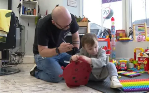 A hairdresser kneel next to a neurodivergent child in a play area in a barber shop