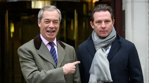 A grinning Nigel Farage points to new Reform UK Treasurer Nick Candy who is wearing a neutral expression. Candy is wearing a scarf and a winter coat, Farage is in a coat over a suit and striped tie