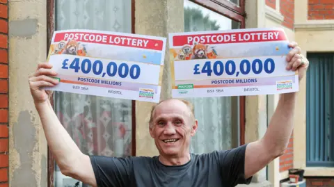 Tony Labruna smiling in front of his house, holding up two postcode lottery 'cheques', both of which say £400,000. 