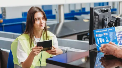 Business Disability Forum A young woman holding a passport wearing a high-vis jacket at an airport. She is looking towards another individual who has their hands on the table of the check-in point. There is a blue poster in the foreground as well as a computer.