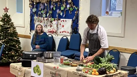 BBC A chef, in a white shirt and a black and white striped apron, cutting an onion. There are different vegetables on the table. There is a Christmas tree to the left and a woman sat on a blue chair waiting blue. 