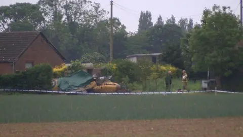 The mangled wreckage of the yellow Spitfire in grassland at the side of a house with two people standing near it and a police cordon. One of those people is a fire officer, who is holding his helmet. 