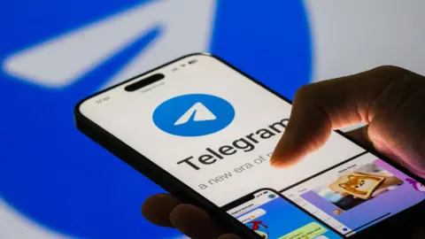 A closeup of an iPhone in a person's hand showing a Telegram screen with the logo, which resembles a stylised paper plane in a blue circle