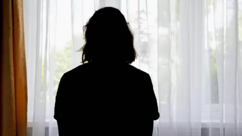 A black silhouette of the back of a woman looking out of a window through white net curtains.