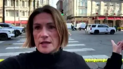 Anna Adams is pointing at a police car, she is stood behind a yellow police tape line in New Orleans