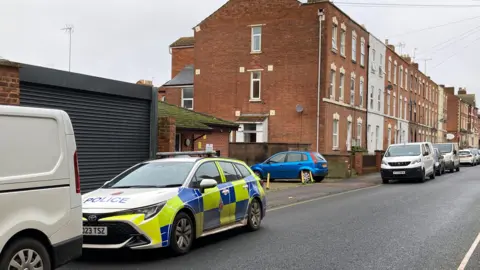 A police car in the foreground of a residential street with tall brick buildings lining the road