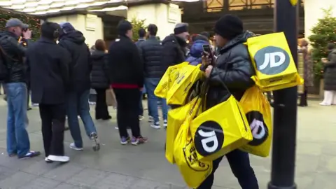 A man with several shopping bags from JD sports walking past a Boxing Day queue in central London's shopping district.