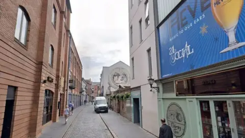 A small street with grey cobblestones and paving. On one side is a red brick building with black shutters. On the other is a number of businesses, the one closest to the camera has a light green front with a large blue billboard. 